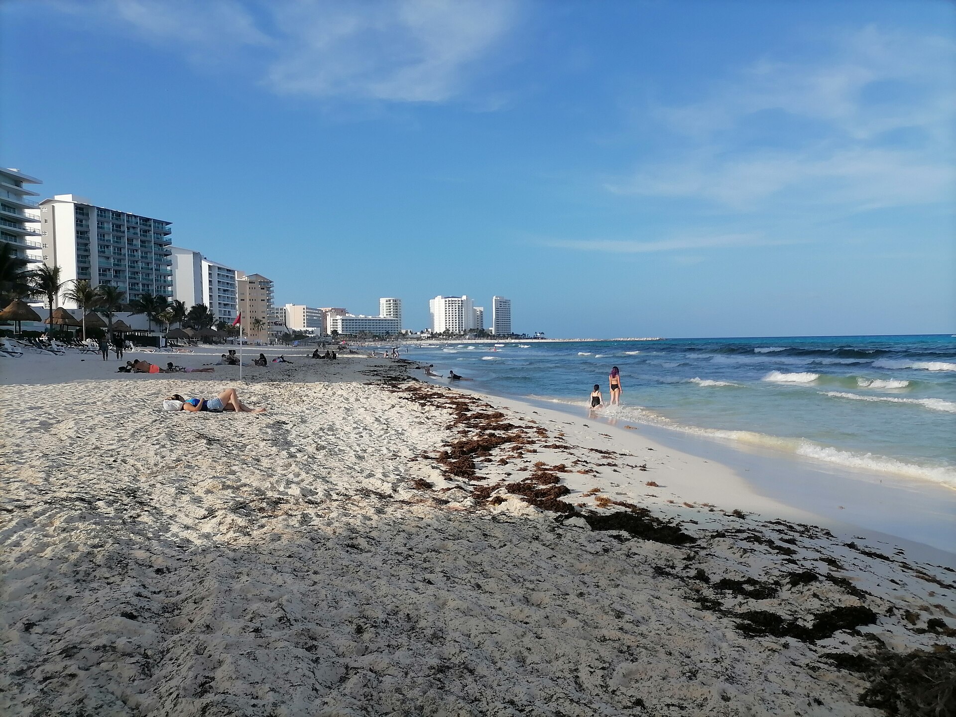 Wide public beach at Playa Delfines in Cancún, with hotels in the distance and some seaweed on the sand